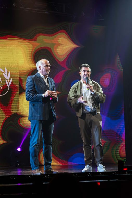 Dos presentadores en un escenario con pantalla de colores detrás